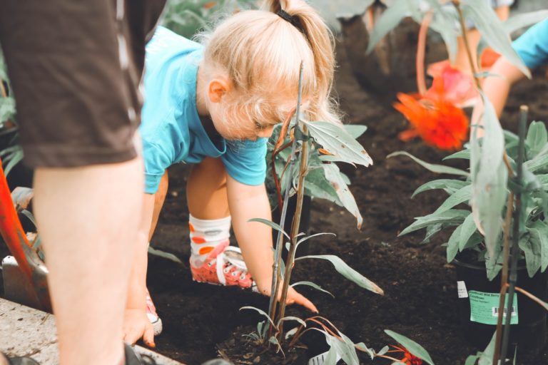 a photoo f a young girl bending over in a garden setting. she has blode hair and is planting a small plant.