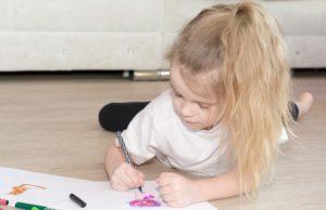 a small girl with blond hair lays on her stomach, hold a pencil and is drawing