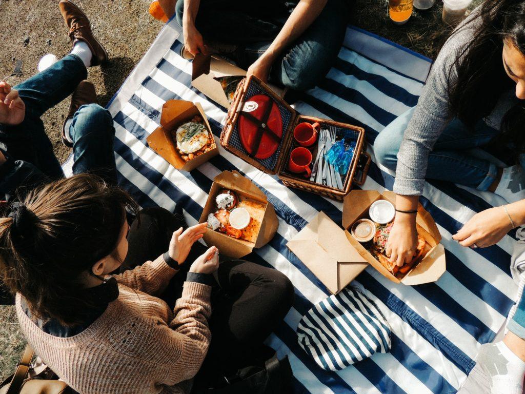 4 people sat around a picnic bbasket on a stripy blanket with breakfast