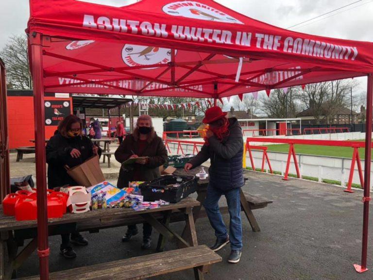 Food bank set up by Ashton United during lockdown. 3 people are under an Ashton United tent, with food parcels for vulnerable people.